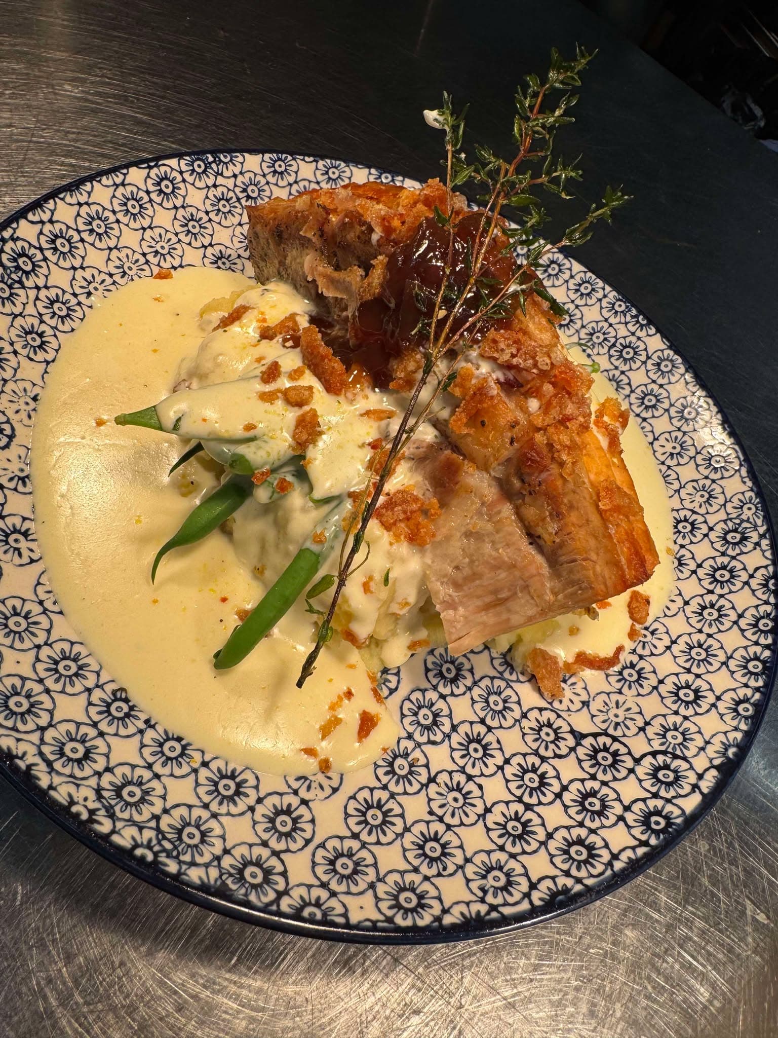 A crisp-skinned pork belly with mash, green beans and a cream sauce on a blue patterned plate, finished with thyme