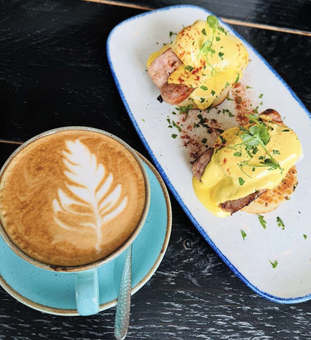 A latte with leaf latte art in a teal cup beside a plate of eggs benedict