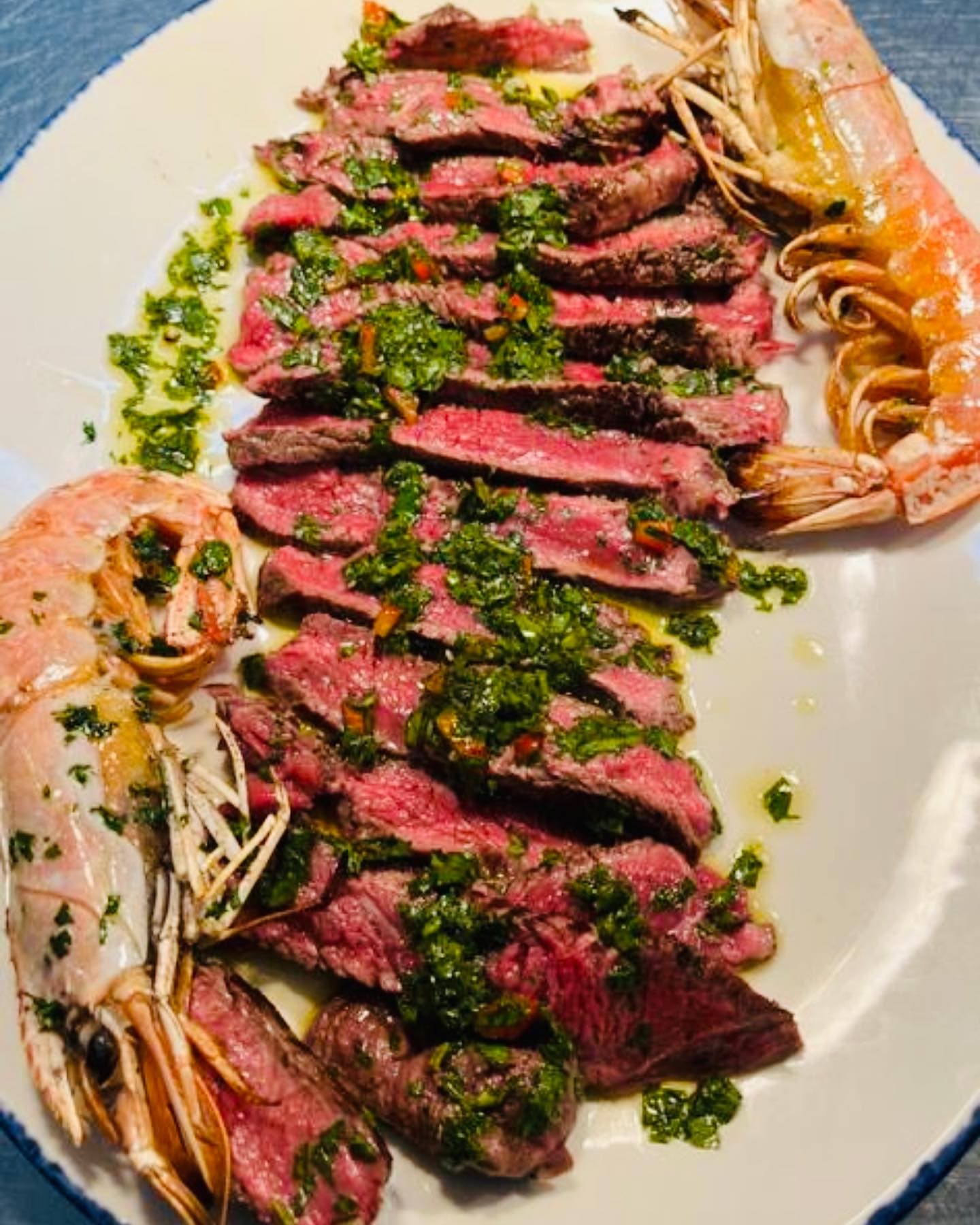 Sliced rare rump steak under chimichurri, flanked by two whole head-on king prawns
