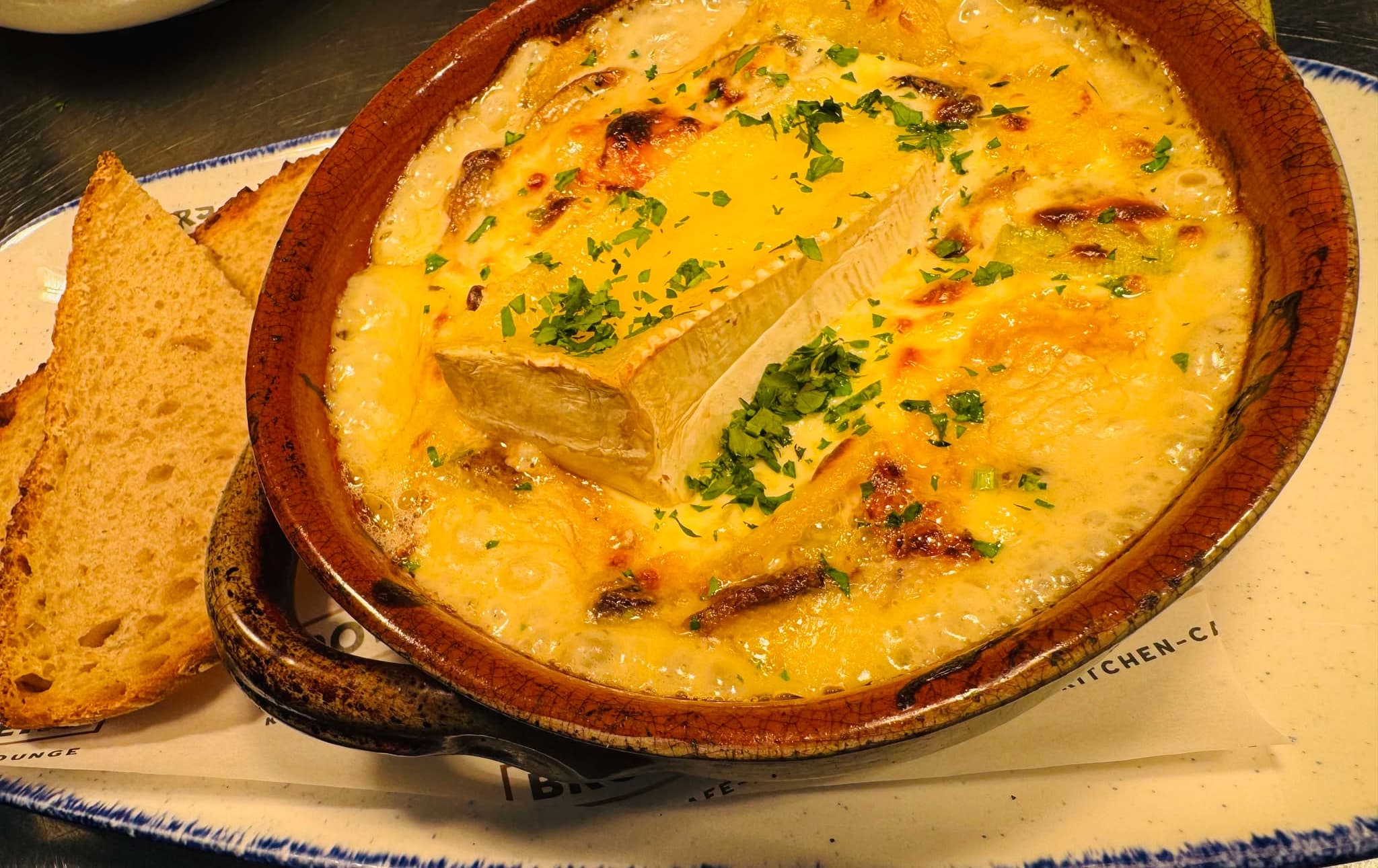 A whole soft cheese baked into a bubbling terracotta pot, scattered with parsley and served with toasted sourdough